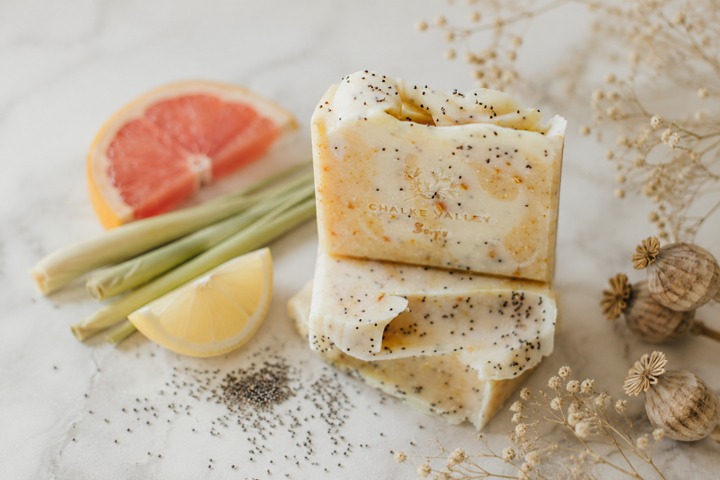Bars of citrus soap with poppy seeds on a surface with lemon, grapefruit, and dried flowers.