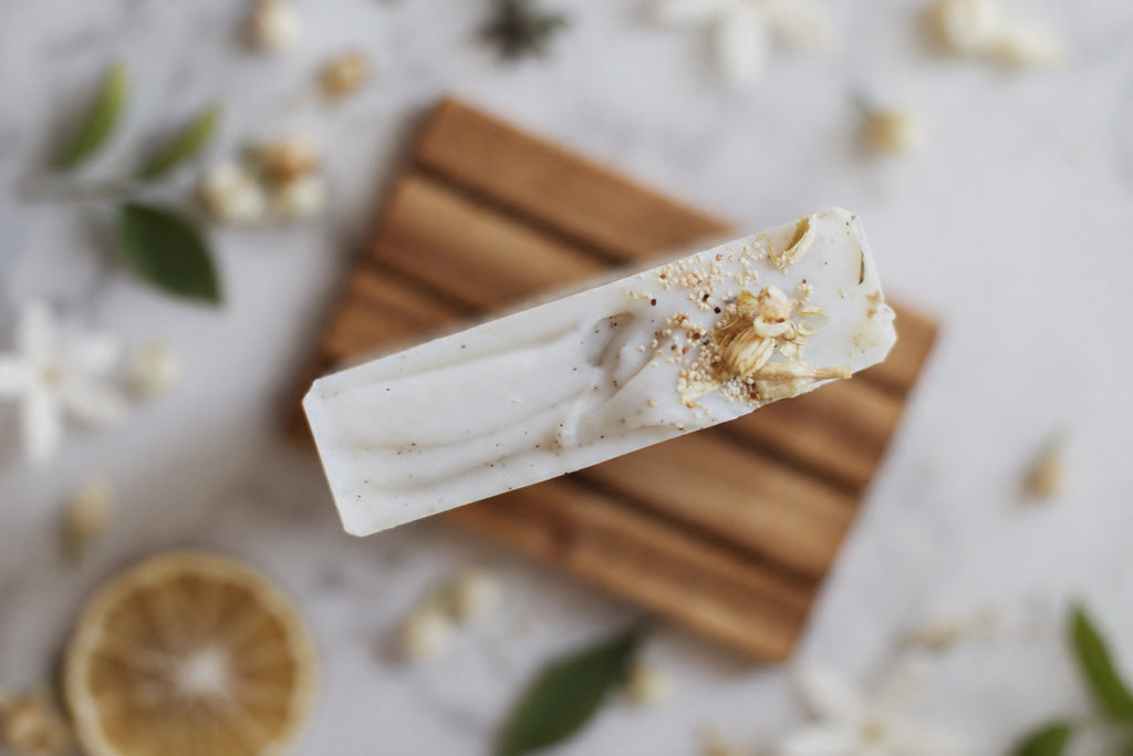 A rectangular bar of white soap with dried botanical inclusions and a sprinkle of brown powder, resting diagonally on a small wooden board with scattered flowers and green leaves around it.